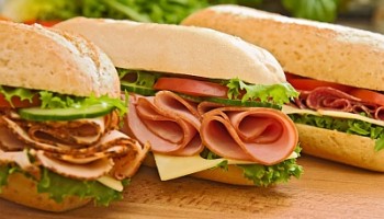Three fresh sub sandwiches - turkey breast, ham & swiss and salami on a cutting board. Focus on the ham sandwich